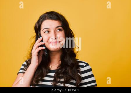 Jeune femme souriant tout en parlant au téléphone, isolée sur fond jaune, l'air pensif Banque D'Images
