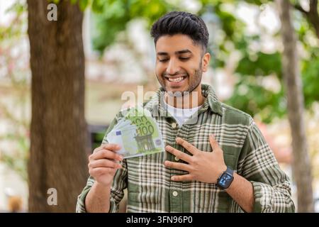 Homme indien tenant de l'argent marchant dans une rue de la ville avec un sourire joyeux célébrant le succès. Touriste arabe montrant des billets en euros en plein air avec le poing levé et une émotion de gagnant heureux après avoir remporté un accord. Banque D'Images