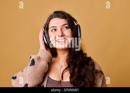 Jeune femme écoutant de la musique avec des écouteurs et souriant contre un fond beige chaud Banque D'Images