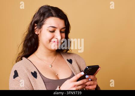 Jeune femme souriante envoyant un message sur son smartphone, portant un cardigan en forme de cœur sur un fond beige chaud Banque D'Images