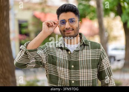 Homme indien mettant ses lunettes en marchant dans une rue de la ville près d'arbres avec un regard portrait concentré. Touriste arabe ajustant ses lunettes en plein air en milieu urbain Banque D'Images