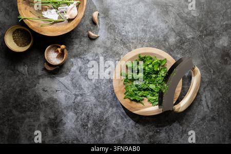 Herbes vertes hachées dans un petit bol en bois avec un couteau mezzaluna et d'autres ingrédients de cuisine sur une surface rustique Banque D'Images