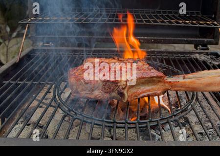 Un épais steak de tomahawk de qualité supérieure se fait griller sur des flammes nues et du charbon de bois chaud sur un barbecue extérieur avec de la fumée qui s'élève. Banque D'Images