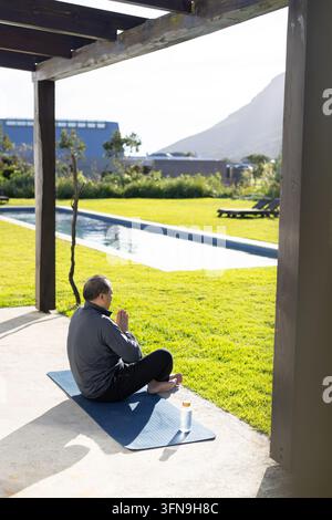 Homme asiatique âgé assis en tailleur portant des vêtements de sport sur un tapis bleu au bord de la piscine, espace de copie Banque D'Images