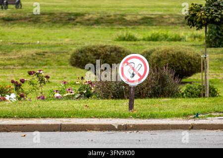 Un panneau rond indiquant une zone non fumeur est fixé sur un pieu en bois le long d'un chemin pavé dans un parc devant des pelouses vertes, des buissons bien entretenus et des parterres de fleurs. Le o Banque D'Images