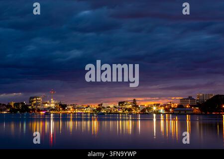 Vue de la skyline de la ville de Tauranga au crépuscule avec des lumières éclatantes se reflétant sur l'eau calme du port sous un ciel dramatique au coucher du soleil à Tauranga, Bay of Plenty Banque D'Images