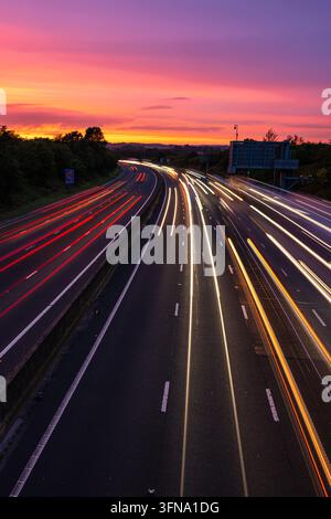 Traînées de feux de circulation à longue exposition sur une autoroute très fréquentée s'incurvant vers un ciel de coucher de soleil violet et orange vif à l'heure de pointe du soir. Banque D'Images