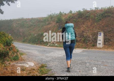 Un pèlerin portant un sac à dos turquoise marche le long d'une route rurale sur le Camino Primitivo, avec un marqueur du Camino de Santiago visible sur le côté, également connu sous le nom de Banque D'Images