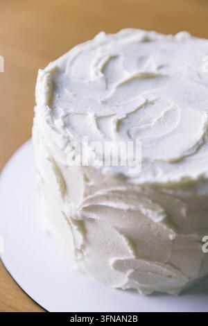 Vue rapprochée d'un gâteau rond fait maison recouvert d'un glaçage blanc texturé, présenté sur une assiette blanche simple posée sur une table en bois. Banque D'Images