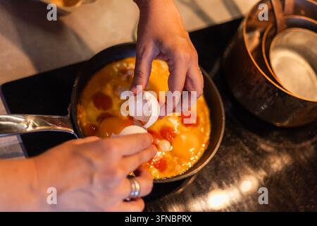 Une paire de mains casse un œuf au-dessus d'une poêle contenant un mélange d'œufs crus. Le gros plan montre les demi-coquilles tenues au-dessus de la poêle sur une cuisinière sombre Banque D'Images