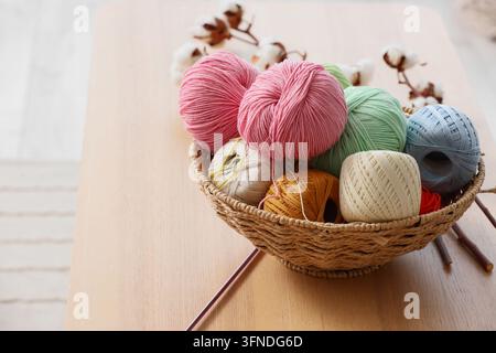 Artisanat et loisirs créatifs, boules de laine colorées dans un panier en osier avec des fleurs en coton pour des projets de tricot et de crochet sur une surface de table en bois Banque D'Images