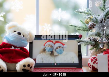 Couple heureux portant un masque médical. Homme et femme se saluant lors d'un appel vidéo. Ordinateur portable sur le rebord de la fenêtre devant une décoration d'arbre de Noël. Vacances de Noël pendant c Banque D'Images