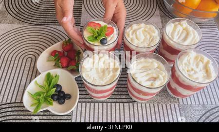 Mains de femme servant une série de desserts tiramisu aux fraises sans œufs faits maison dans des verres, garnis de baies de menthe fraîche et d'une couche de crème mascarpone. Banque D'Images