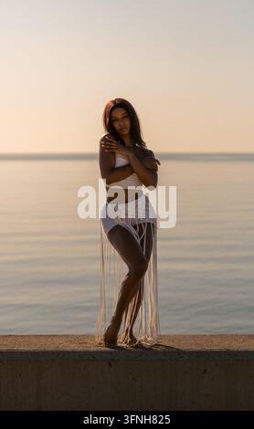 Une belle Cubaine se tient sur un mur au bord de la mer, s'enlaçant avec les bras croisés et arborant une expression sereine et pensive ; elle est vêtue d'un Banque D'Images