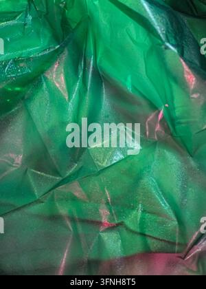 Vue abstraite en gros plan d'un revêtement plastique vert froissé avec des nuances roses et rouges subtiles. Texture de surface réfléchissante avec un éclairage doux, suggérant Banque D'Images