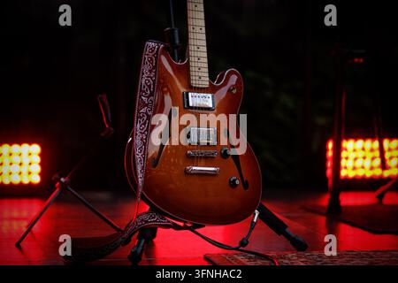 Vue avant d'une guitare électrique semi-cavité brune sur un support avec une sangle décorée sous un éclairage rouge des projecteurs. Banque D'Images