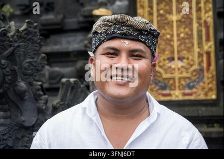 Portrait d'un homme balinais souriant portant des couvre-chefs traditionnels à motifs et une chemise blanche, debout devant des sculptures ornées de temples hindous et un doré Banque D'Images