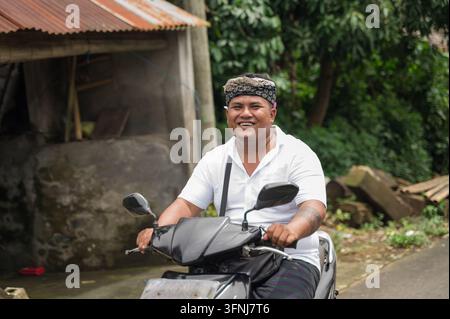 Homme balinais souriant portant un foulard traditionnel udeng conduit une scooter le long d'une route de village rural à Bali, Indonésie, entouré d'une végétation tropicale Banque D'Images