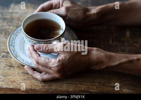 Mains humaines tenant une tasse à thé en porcelaine vintage remplie de thé noir chaud sur une table en bois rustique sombre Banque D'Images