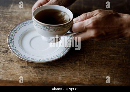 Mains humaines tenant une tasse à thé en porcelaine vintage remplie de thé noir chaud sur une table en bois rustique sombre Banque D'Images