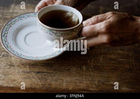 Mains humaines tenant une tasse à thé en porcelaine vintage remplie de thé noir chaud sur une table en bois rustique sombre Banque D'Images