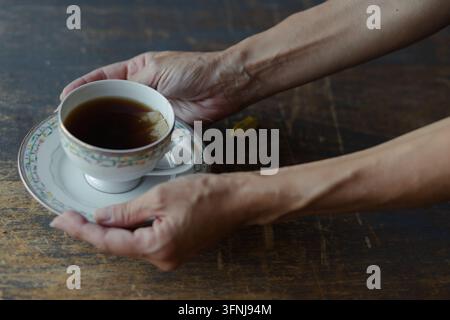 Mains humaines tenant une tasse à thé en porcelaine vintage remplie de thé noir chaud sur une table en bois rustique sombre Banque D'Images