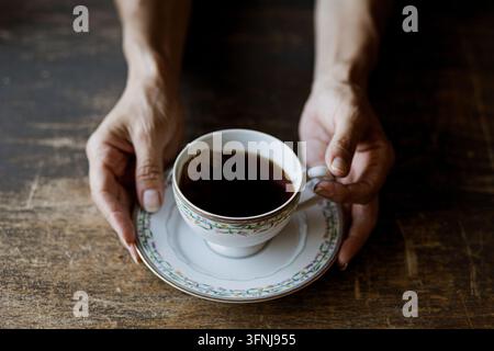 Mains humaines tenant une tasse à thé en porcelaine vintage remplie de thé noir chaud sur une table en bois rustique sombre Banque D'Images