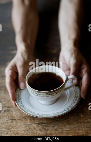 Mains humaines tenant une tasse à thé en porcelaine vintage remplie de thé noir chaud sur une table en bois rustique sombre Banque D'Images
