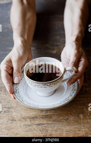 Mains humaines tenant une tasse à thé en porcelaine vintage remplie de thé noir chaud sur une table en bois rustique sombre Banque D'Images