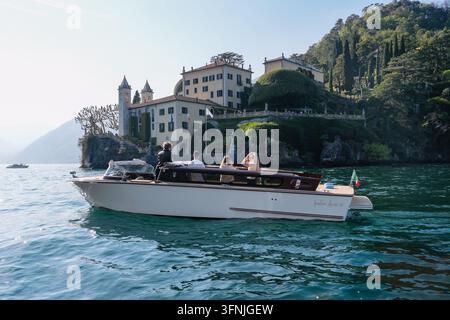 Varenna, Italie - avril, 05, 2026 : Un bateau à moteur blanc moderne transportant un groupe de touristes glisse sur le lac bleu clair devant la villa historique italienne Banque D'Images