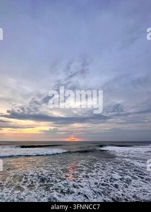 Le soleil couchant brille au-dessus des vagues écumantes de l'océan sous des nuages vespéraux superposés. Banque D'Images