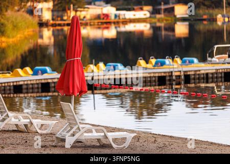 Une vue extérieure d'une plage de station balnéaire vide au bord du lac avec des chaises longues en plastique blanc et un parasol rouge plié sur le sable, avec un arrière-plan flou d'un Banque D'Images
