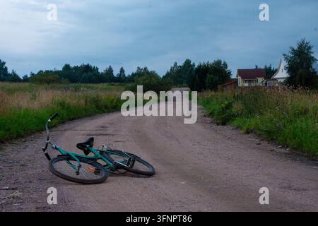 Photographie atmosphérique de la campagne montrant un vélo vert gisant sur le côté sur une route de terre sinueuse. La route mène vers de petites maisons de village et gr Banque D'Images