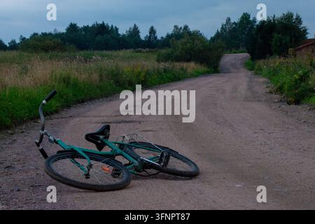 Photographie atmosphérique de la campagne montrant un vélo vert gisant sur le côté sur une route de terre sinueuse. La route mène vers de petites maisons de village et gr Banque D'Images
