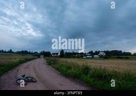 Photographie atmosphérique de la campagne montrant un vélo vert gisant sur le côté sur une route de terre sinueuse. La route mène vers de petites maisons de village et gr Banque D'Images