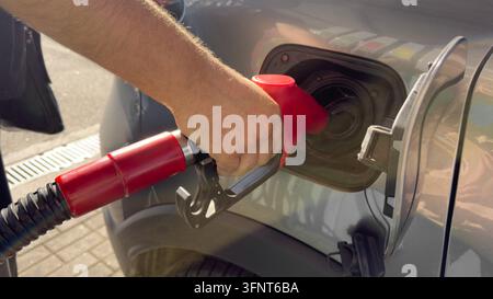 Gros plan d'une main de conducteur tenant un pistolet de carburant rouge et l'insérant dans le réservoir d'essence d'une voiture grise à une station-service extérieure. Concept de ravitaillement de véhicule Banque D'Images