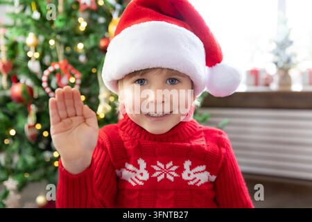 Enfant joyeux portant un chapeau du Père Noël. Enfant jouant à la maison. Portrait d'enfant à Noël. Enfant drôle saluant dans un appel vidéo. Concept de fête de Noël Banque D'Images
