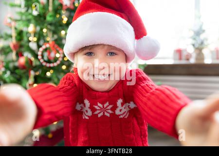 Enfant joyeux portant un chapeau du Père Noël. Enfant jouant à la maison. Portrait d'enfant à Noël. Enfant drôle saluant dans un appel vidéo. Concept de fête de Noël Banque D'Images