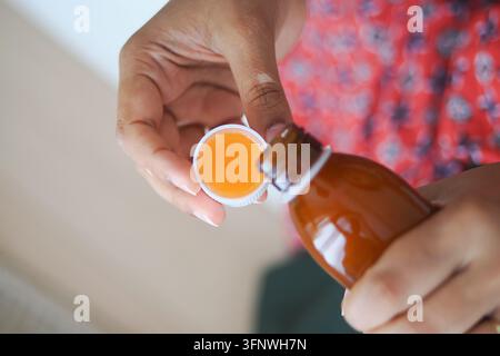 Person versant un médicament liquide d'une bouteille dans un petit bouchon dans un cadre domestique Banque D'Images