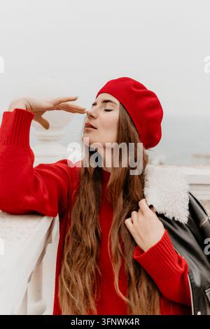 Portrait d'une femme portant un béret rouge, posant en plein air dans un pull élégant près d'une rambarde blanche par une journée nuageuse Banque D'Images