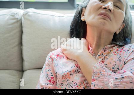 Femme asiatique âgée souffrant de douleurs sévères à l'épaule et au cou, tenant son épaule avec une expression douloureuse tout en étant assise sur un canapé à la maison Banque D'Images