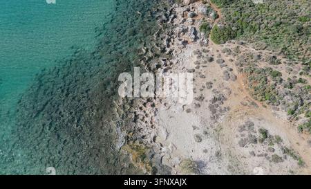 Vue aérienne de dessus d'une côte rocheuse rencontrant une eau de mer turquoise claire, révélant des motifs du fond marin et la végétation côtière en Grèce comme fond Banque D'Images