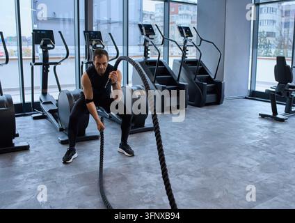 Homme mûr s'entraînant avec des cordes de combat lourdes dans une salle de fitness moderne avec des machines de cardio et de grandes fenêtres Banque D'Images