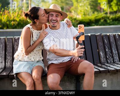 Homme et femme souriants en train d'enregistrer un vlog amusant ensemble dans un parc ensoleillé. La femme embrasse un homme. Banque D'Images