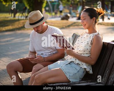 Femme profitant d’un bon livre assise à côté d’un homme utilisant son smartphone sur un banc de parc. Banque D'Images