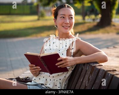 Une femme asiatique d'âge moyen heureuse se détend dans un parc en été, elle lit un livre. Banque D'Images