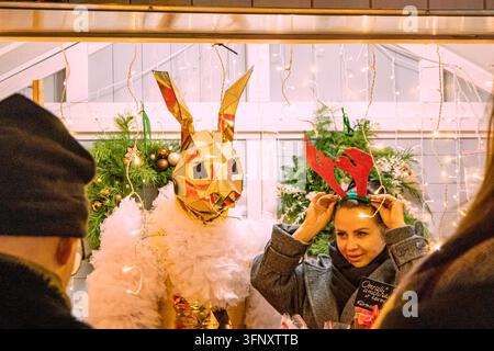 Bialystok, Pologne – 4 décembre 2025 : Une femme essaie des bois de renne à un stand de marché de Noël avec une artiste en costume de lapin doré et une guirlande lumineuse Banque D'Images