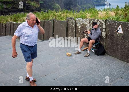 Un touriste passant devant un musicien de rue jouant de la flûte aux falaises de Moher, dans le comté de Clare, en Irlande Banque D'Images
