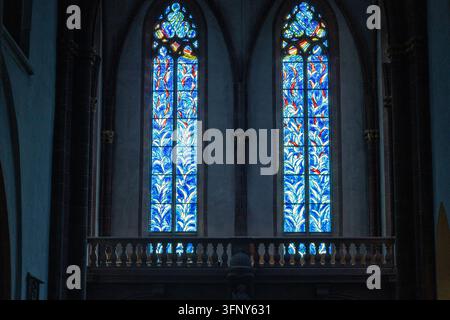 Vue intérieure d'une église historique présentant deux hautes baies vitrales colorées, des colonnes en pierre sombre, des arcs pointus et un balustradier en pierre décoratif Banque D'Images