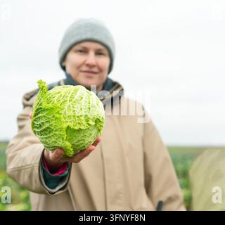 Femme agricultrice fière expose du chou fraîchement récolté dans son jardin, rayonnant de satisfaction avec ses produits cultivés à la maison. Cette jardinière se tient dans le champ, célébrant la joie de cultiver des légumes frais Banque D'Images